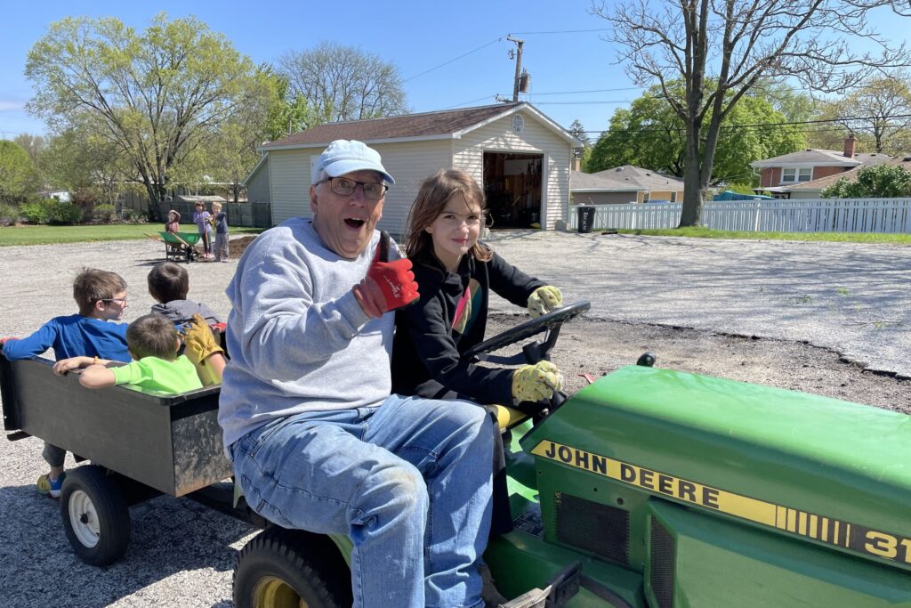 Young girl drives tractor over a parking lot while man gives a thumbs up.