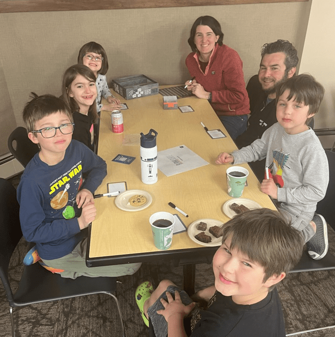 Three boys, two girls, and a man and a woman smiling around a game table.