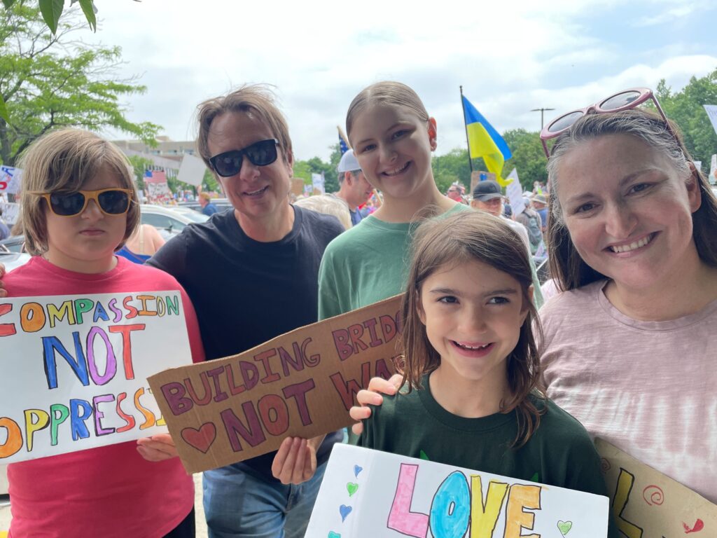 A family holds signs saying "Compassion, not oppression" and "Building bridges, not walls" at a No Kings Protest. 