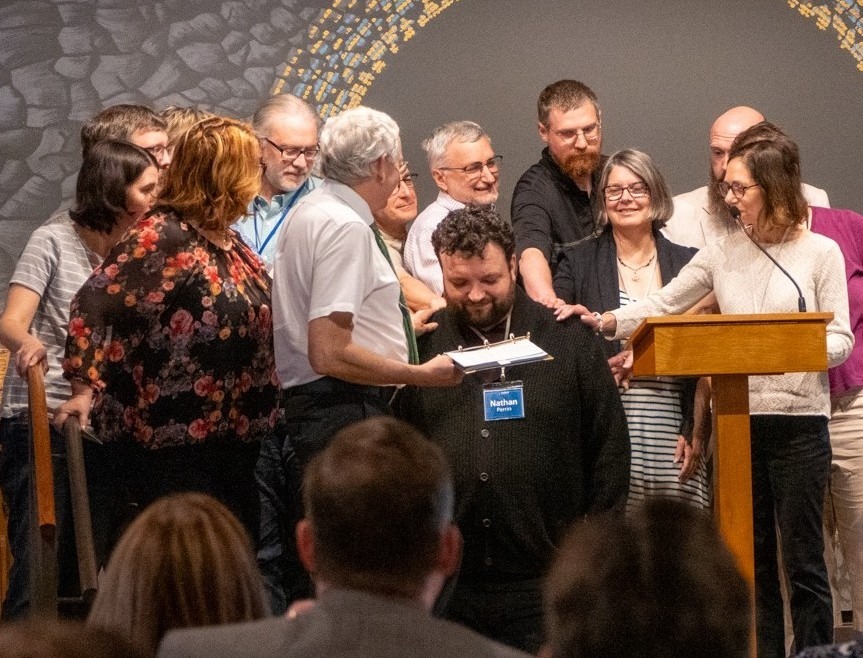 Group of men and women surround a kneeling man at his ordination inside a church sanctuary. 