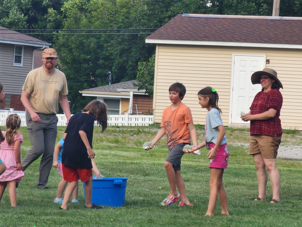 Kids and two adults pick up water balloons from a blue bucket to throw at an outdoor event. 