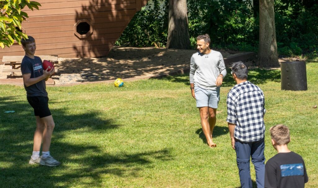 Two teens and two men play catch with a football on a green lawn.