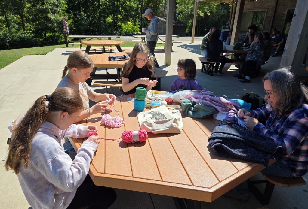 Young teens and a child sit around a picnic table and crochet on a patio. 