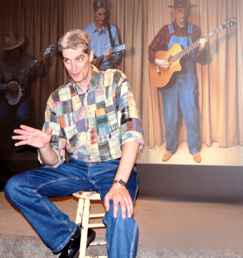 An actor sits on a stool, with a band playing on a video screen behind him.