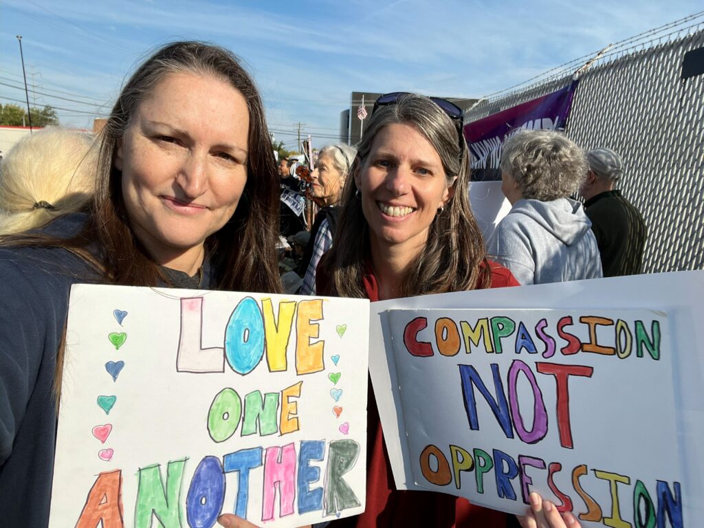 Two women at a peace vigil outside a fenced facility, holding signs reading "Love one another" and "Compassion not oppression." 