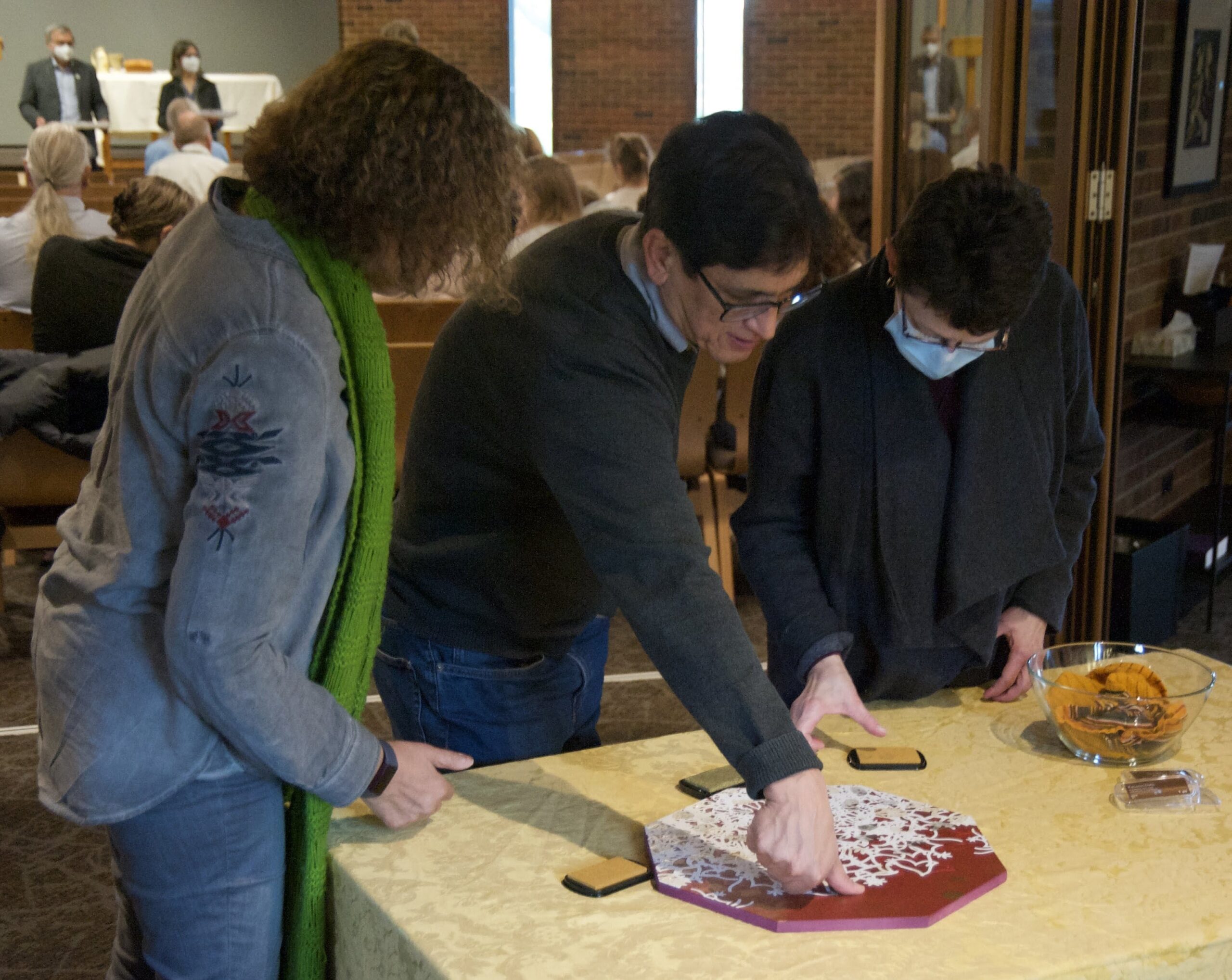 A man surrounded by two women stamps his thumbprint on an octagonal art piece outside a church sanctuary. 