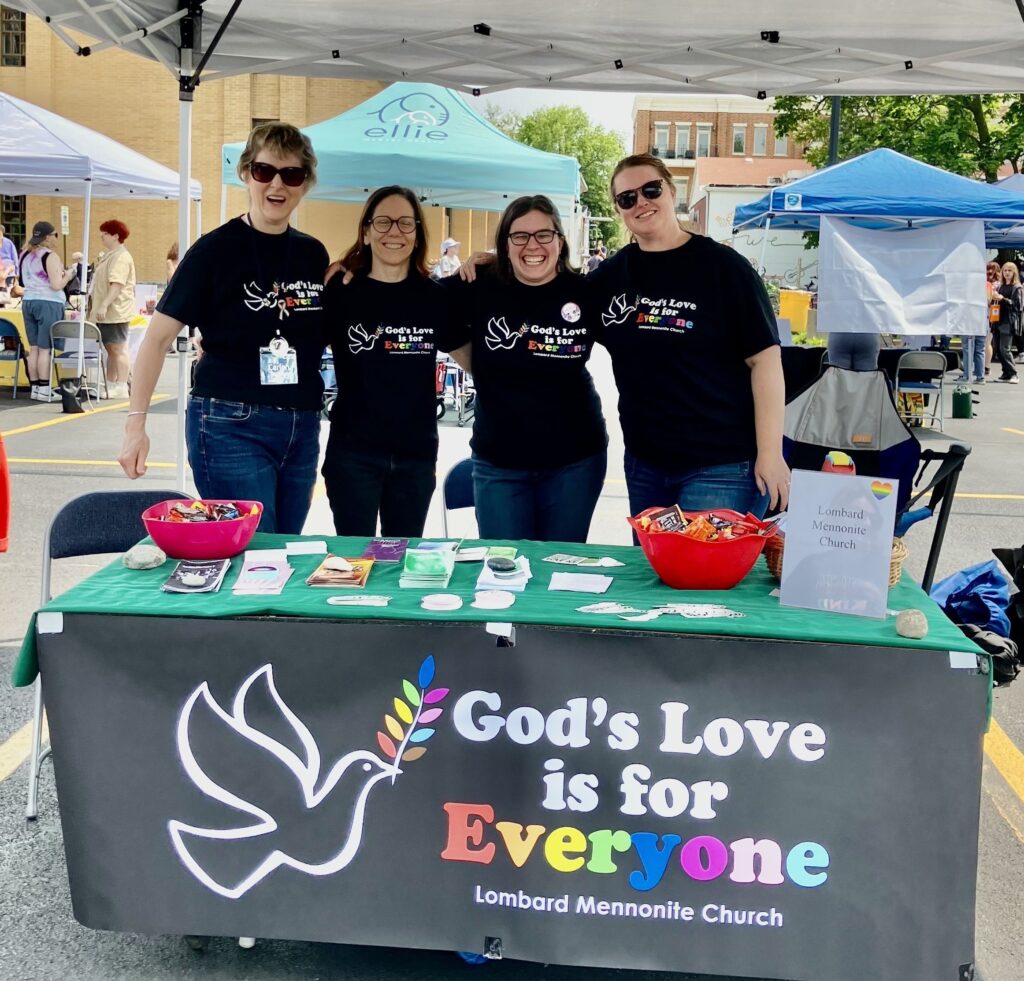 Four women stand at a table with a banner sign: God's Love is for Everyone, Lombard Mennonite Church