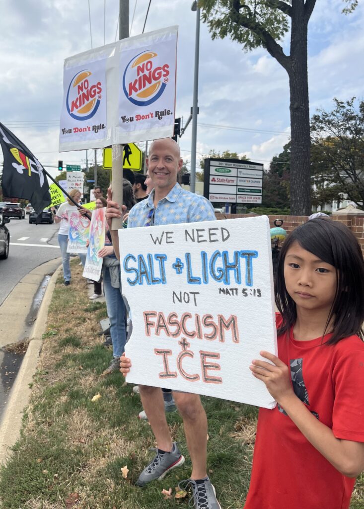 Man with a sign reading, "No Kings; You don't rule." and boy with a sign reading, "We need salt & light (Matt 5:13) not fascism & ice" on the side of an intersection. 