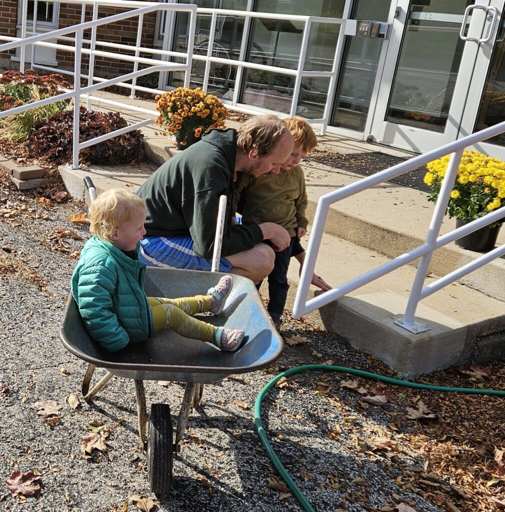 A man, a young boy, and a toddler sitting in a wheelbarrow at a church workday.