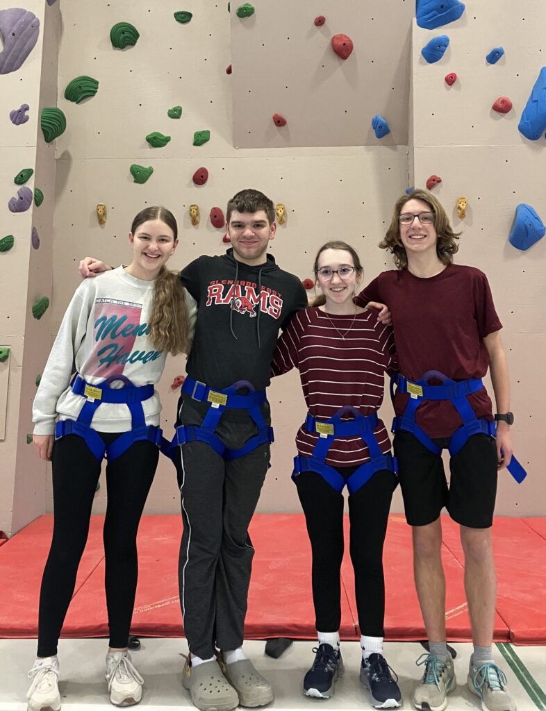 Four teenagers (two boys, two girls) stand in front of a climbing wall. 
