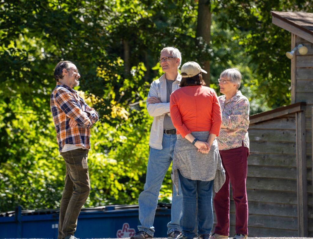 A group of two men and two women chat outside a cabin amid lush greenery. 