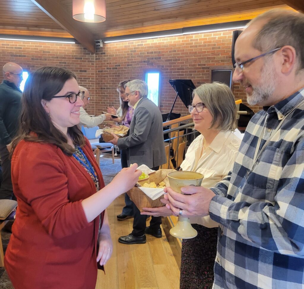 A woman receives bread and juice from two people during a communion service in a church with several others participating in the background.