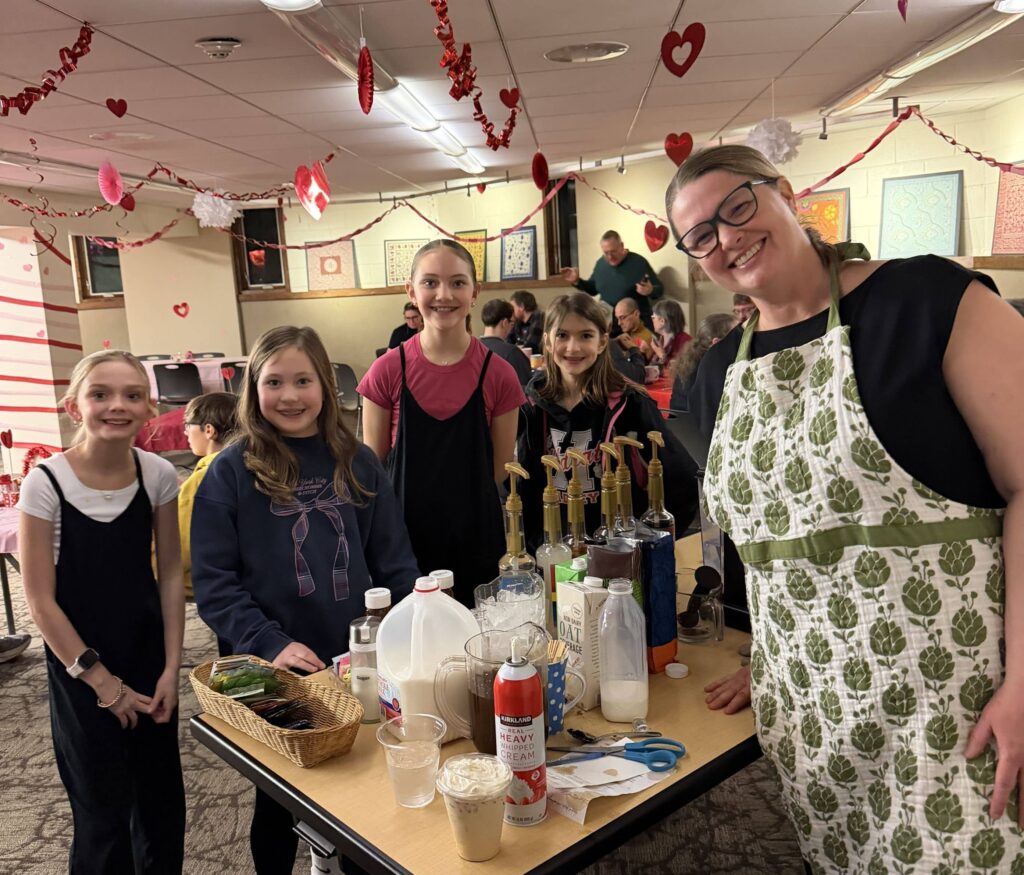A woman and four girls smile around a table containing ingredients to make specialty coffee drinks.