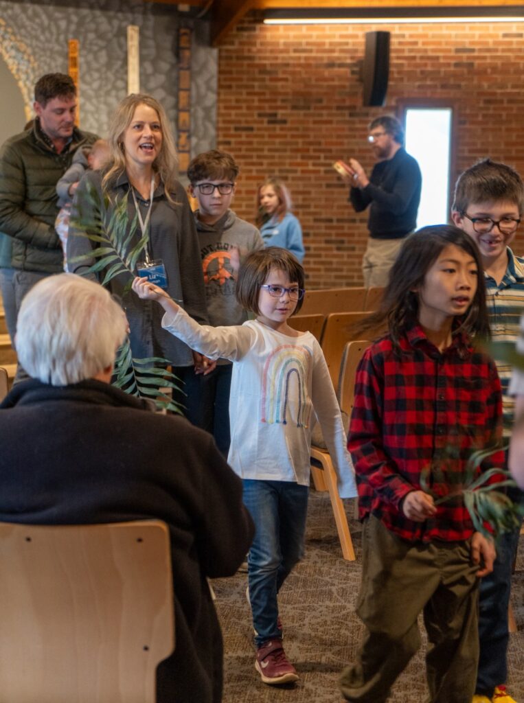 Children and a couple adults wave palm branches as they walk around a church sanctuary.