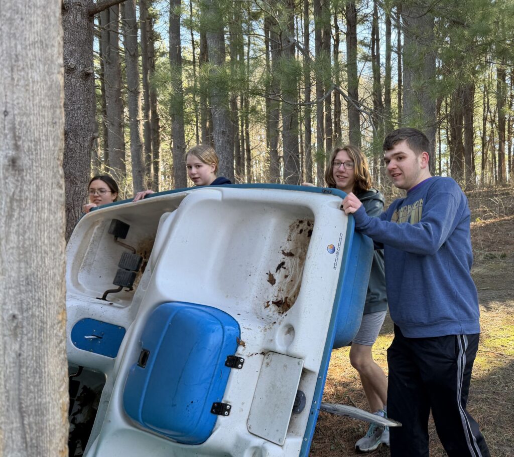A group of four teens push a paddleboat up against a cabin in the woods.