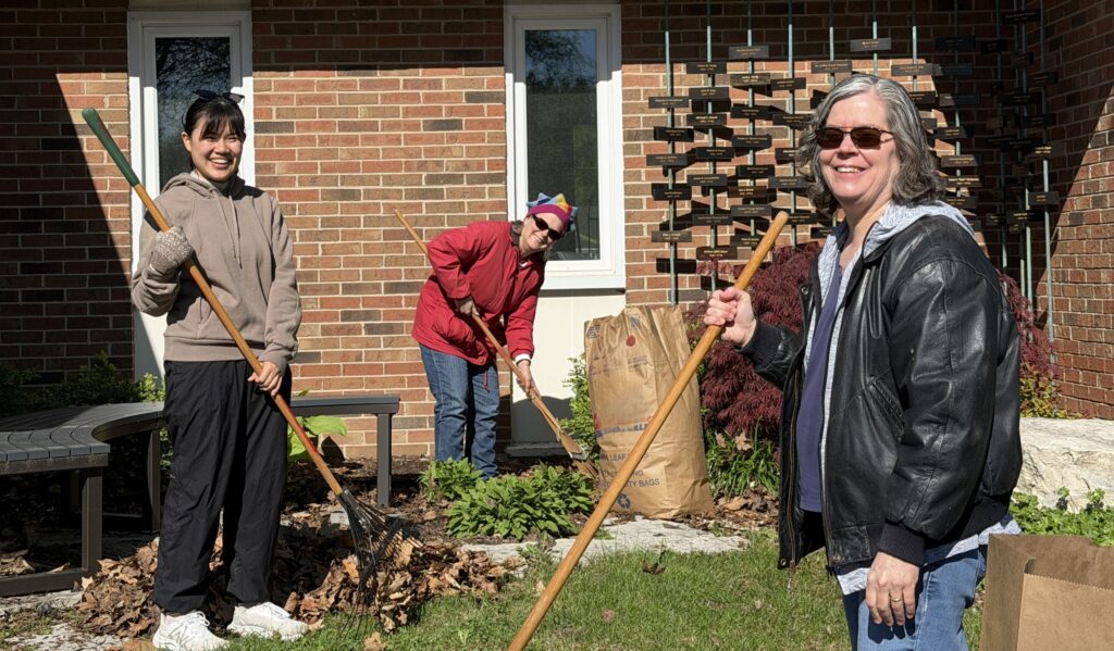 Three women smile as they rake leaves in front of a church wing. 