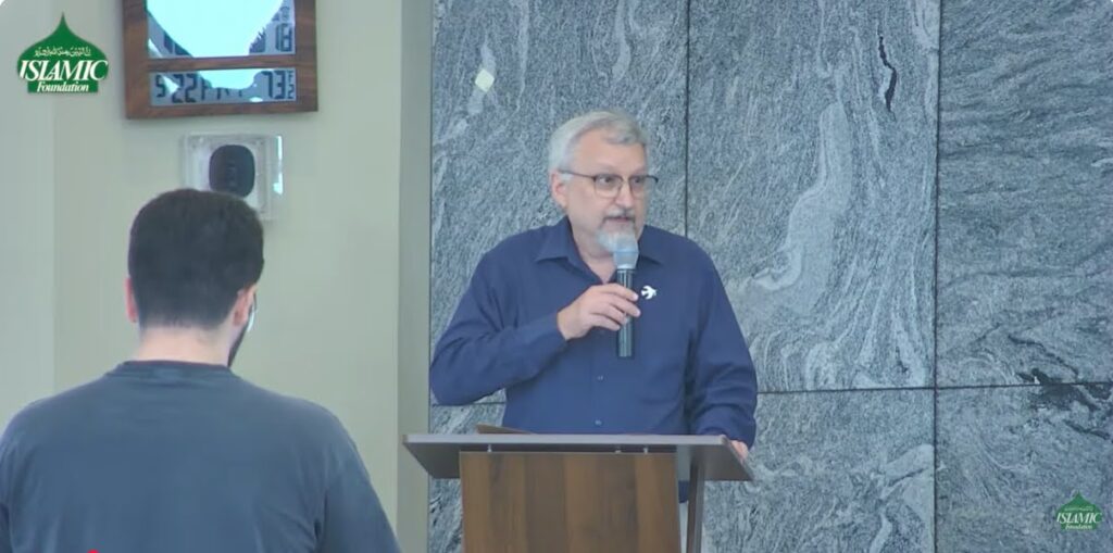 A man holds a microphone and speaks in front of a gathering. 