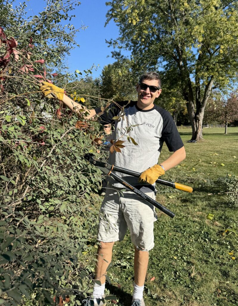 A man with clippers smiles as he trims an overgrown shrub. 