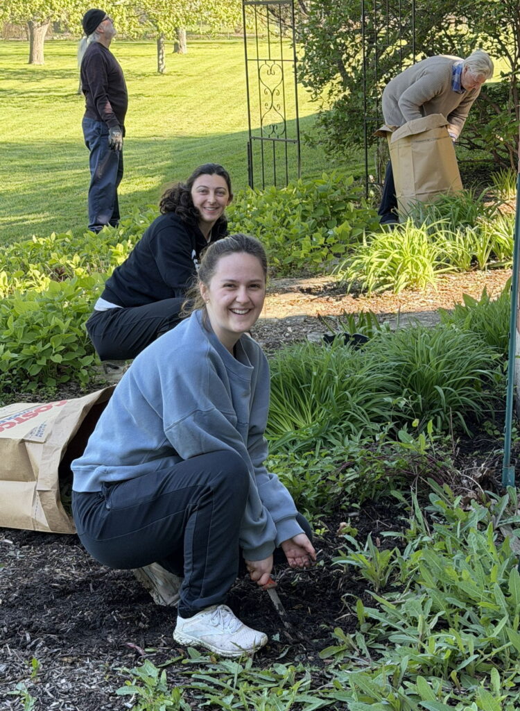 Two young women smile as they pull weeds from landscaping around a church. 