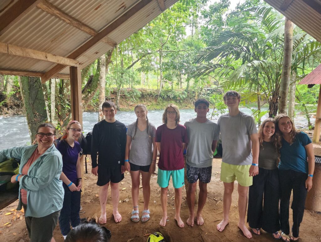 A group of teenagers and three young adults stand inside an open structure in front of a river in the jungle in Costa Rica.