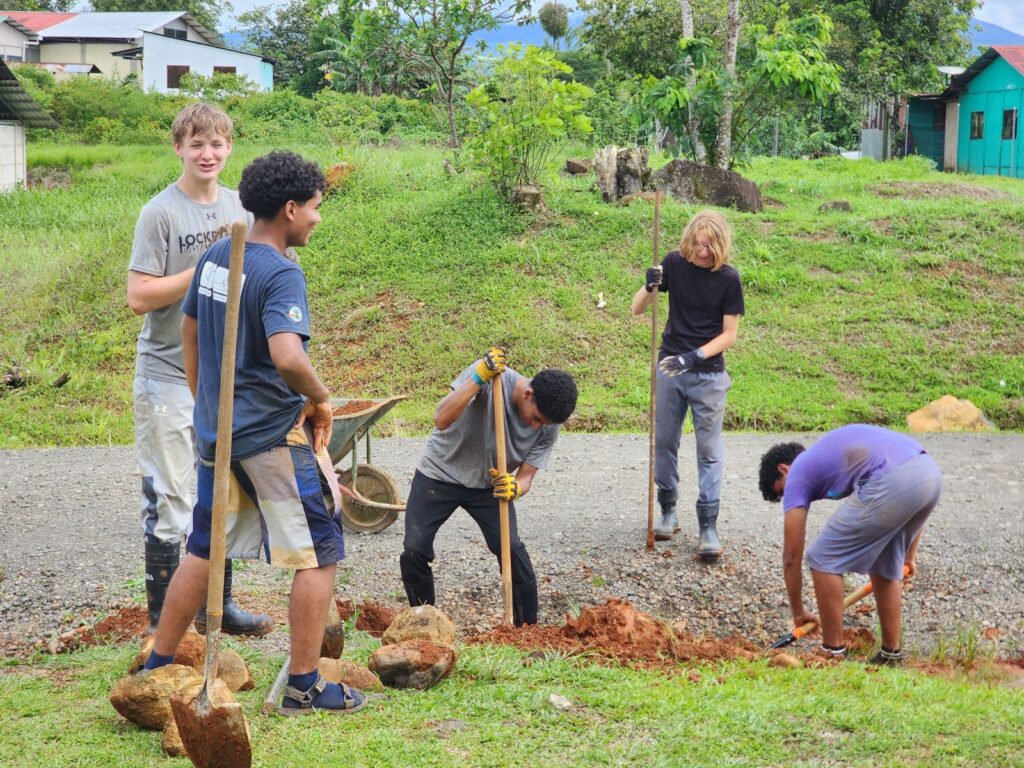 Five teenagers dig a ditch in a green, rural setting.