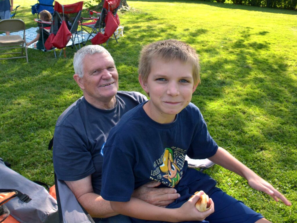 A young boy sits on his grandfather's lap during an outdoor event.