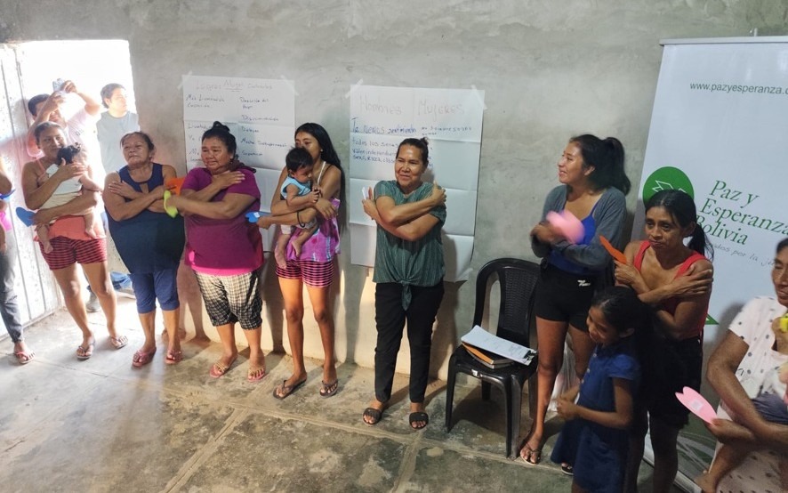 A group of women stand in a circle around the edges of a cement room and hug themselves.