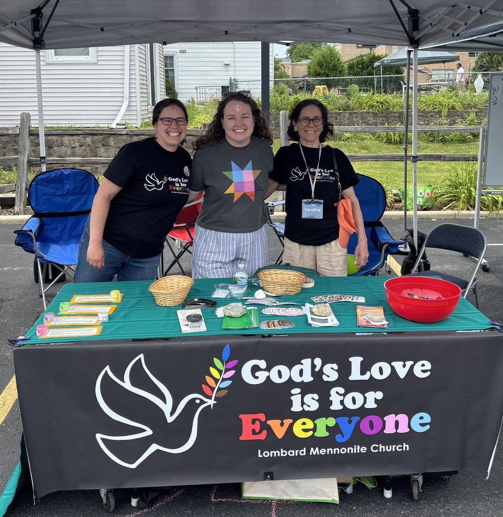 Three smiling women behind a table with a banner that says "God's love is for everyone."