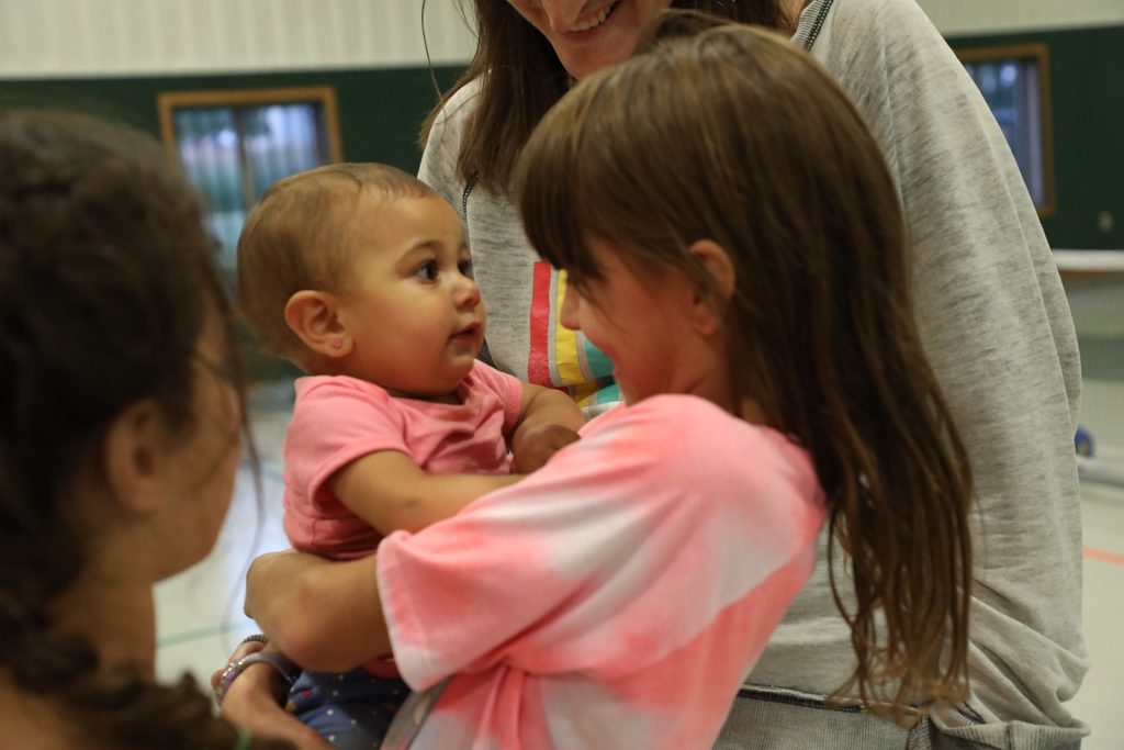 A young girl beams while she holds a baby, surrounded by smiling people.