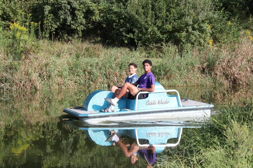 Two boys on a paddle boat on a lake surrounded by vegetation, which reflects in the water.