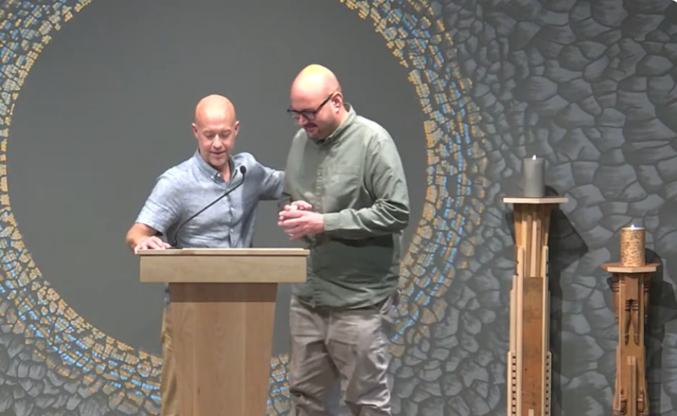 A man introduces another man at a pulpit inside a church sanctuary.