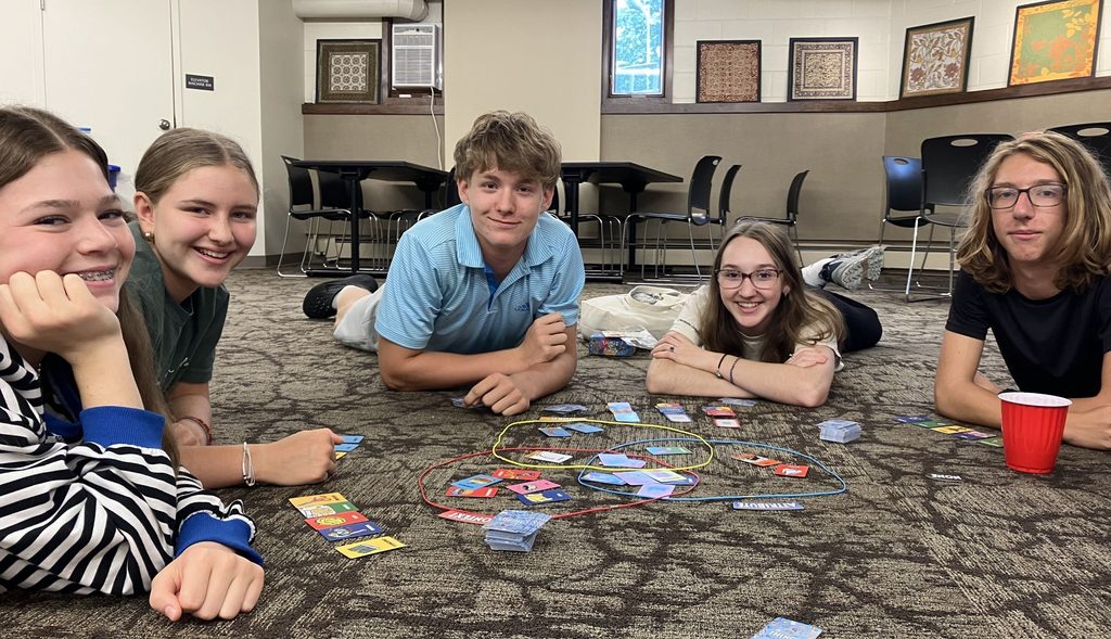 A group of smiling teens lay on the floor around a card game.