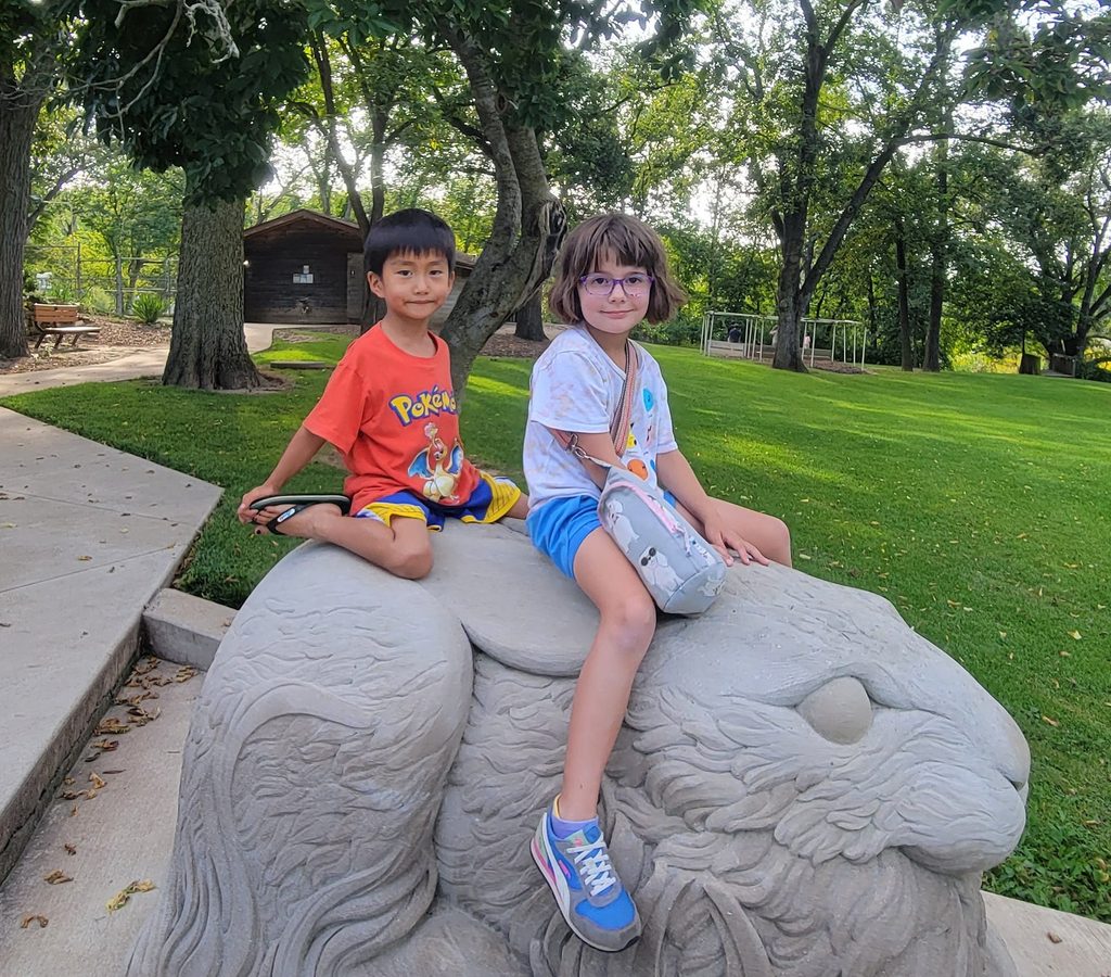 Two children sit on a stone rabbit sculpture at a camp.