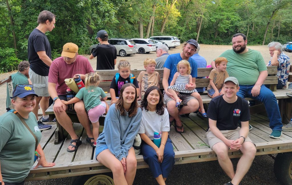 A group of adults and children sit on a hayrack within a wooded camp