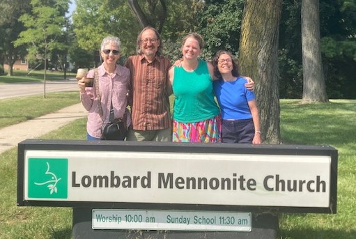 Three women and a man stand behind a church sign that says, "Lombard Mennonite Church."
