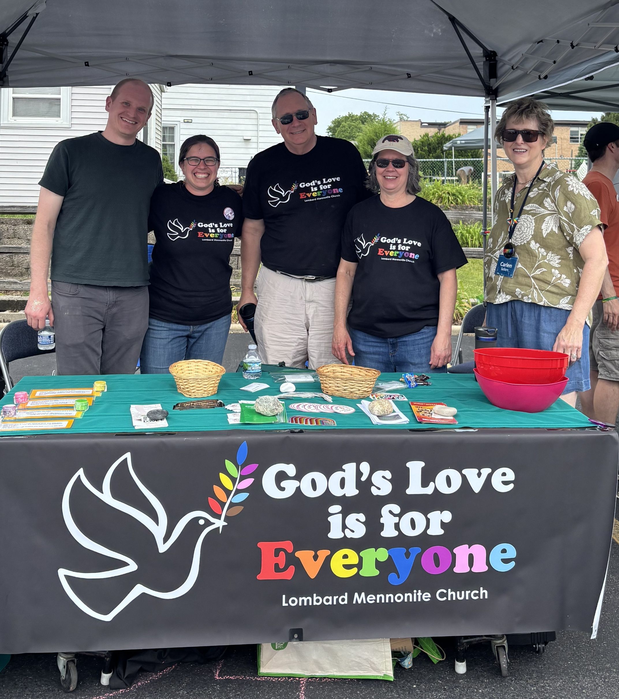 Two men and three women stand behind a table with a banner that says, "God's Love is For Everyone; Lombard Mennonite Church"